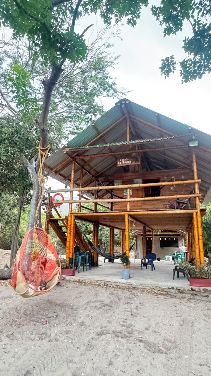 A wooden structure with a covered roof, possibly a cabin or a small building, surrounded by trees and a sandy ground.