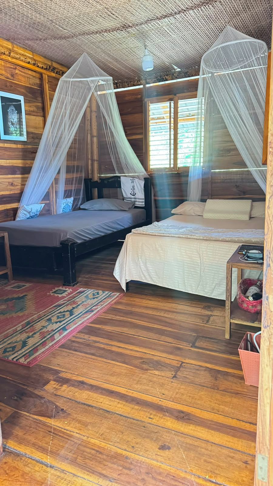 Un dormitorio de cabaña de madera con dos camas, una mesita de noche, una pequeña mesa y una ventana con persianas.