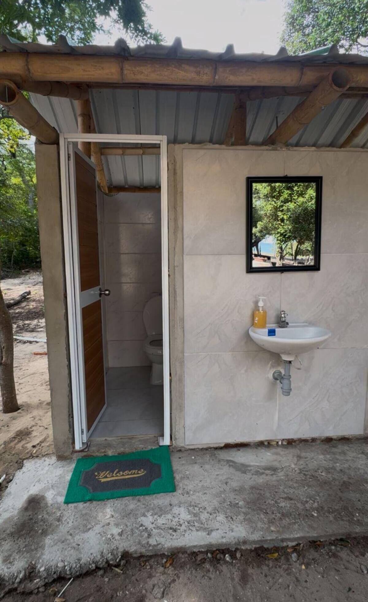 Una pequeña estructura de una sola habitación con un inodoro y un lavabo, un felpudo y un espejo en un marco en la pared.