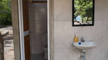 Una pequeña estructura de una sola habitación con un inodoro y un lavabo, un felpudo y un espejo en un marco en la pared.