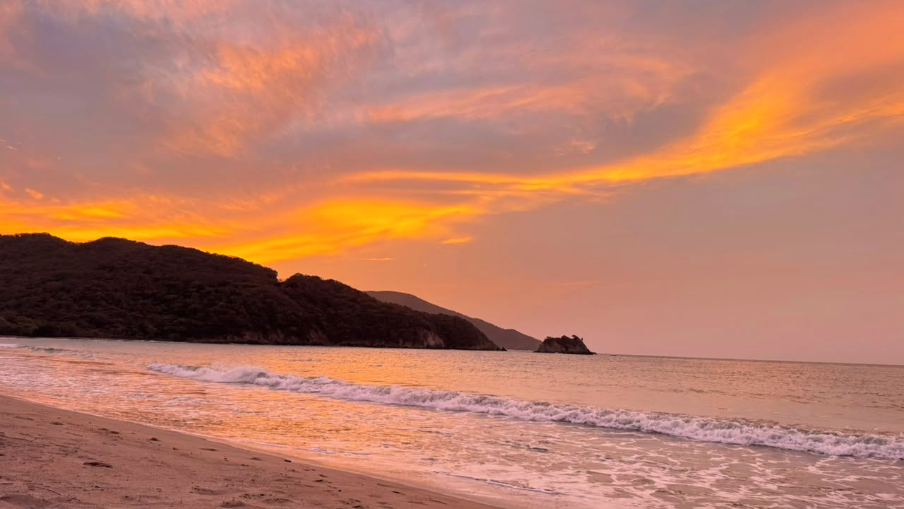 A beach with waves, a sunset, and distant hills.