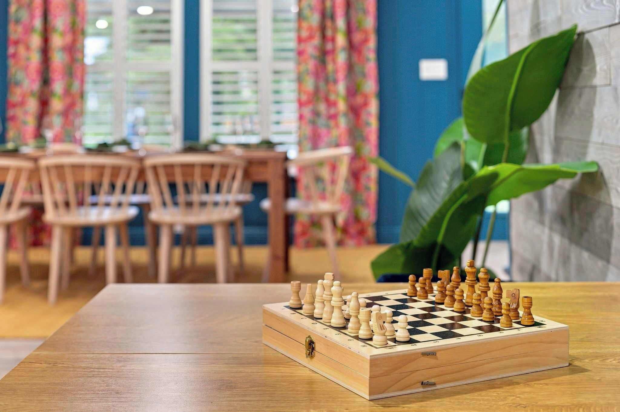 Un échiquier posé sur une table en bois, dans une pièce aux murs bleus et aux rideaux à motifs floraux.