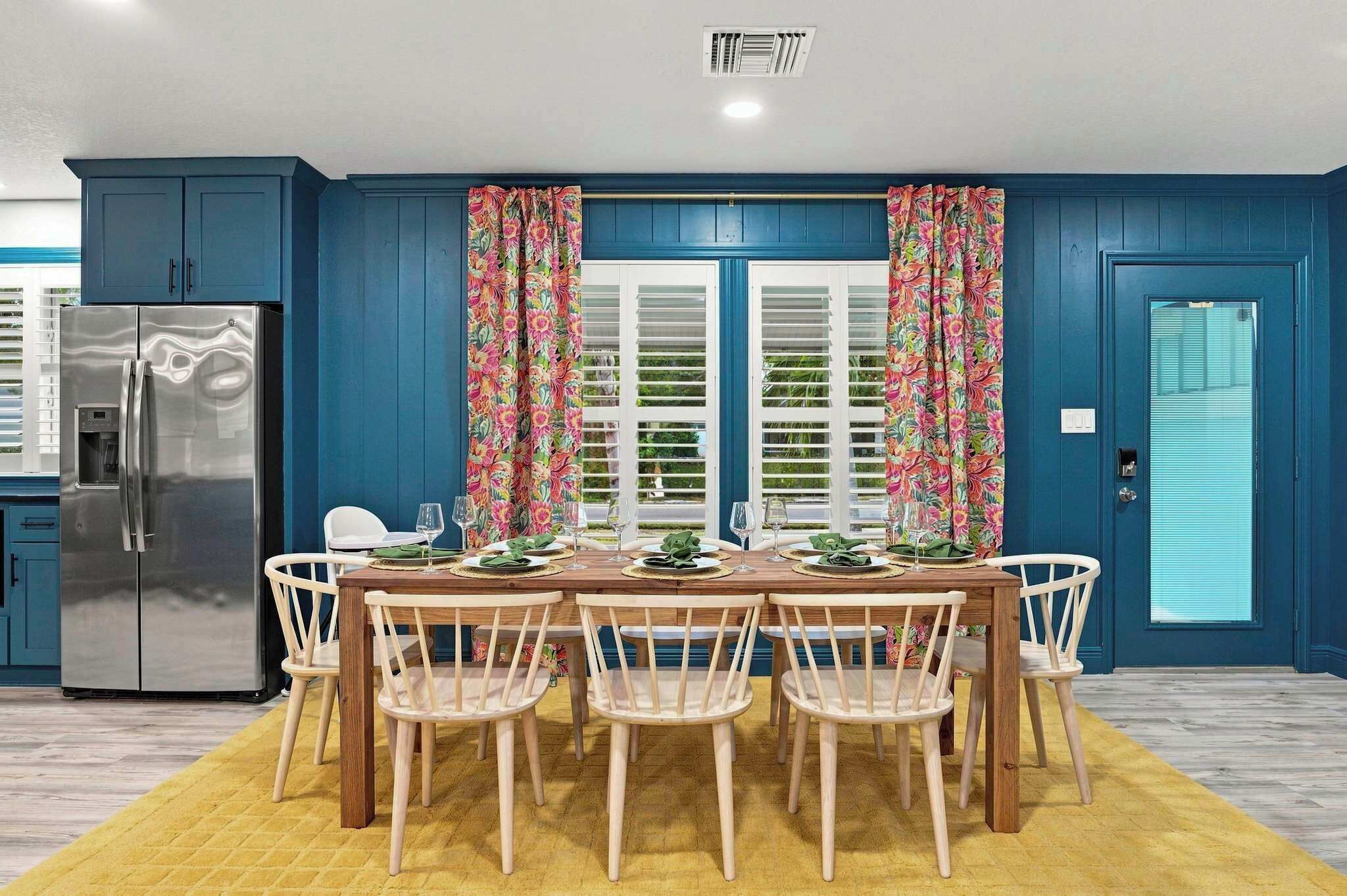 A dining area with a wooden table set for four, surrounded by chairs, in a room with blue walls and a yellow rug.