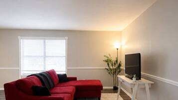 A living room with a red sectional sofa, a small round table, a TV on a white stand, and a floor lamp.