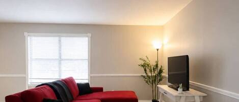 Una sala de estar con un sofá seccional rojo, una mesita redonda, un televisor sobre un mueble blanco y una lámpara de pie.