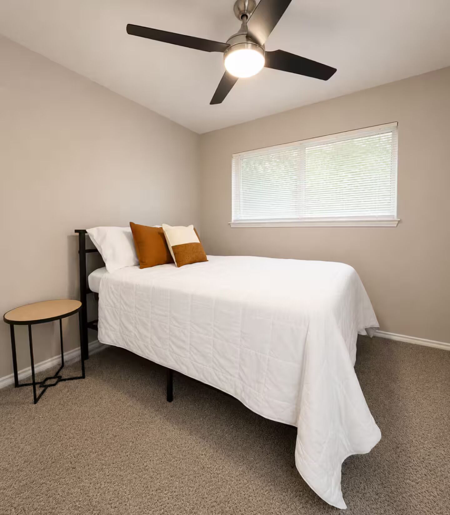 A bedroom with a bed, a ceiling fan, a window with blinds, and a small table.