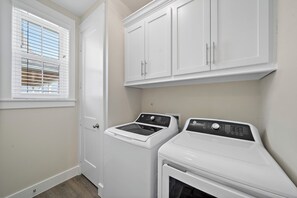 A laundry room with a washer and dryer, white cabinets above, and a window with blinds. - Casa de Miramar 4BR/3BA with 3 King Beds and Beach, Pool & Fishing Pier Access (Galveston)
