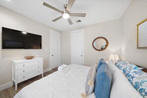Un dormitorio con cama, cómoda, televisor, ventilador de techo, espejo y un cuadro en la pared.