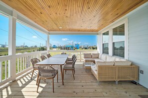 A covered outdoor deck with a table and chairs, a sofa, and a view of the surrounding area. - Casa de Miramar 4BR/3BA with 3 King Beds and Beach, Pool & Fishing Pier Access (Galveston)