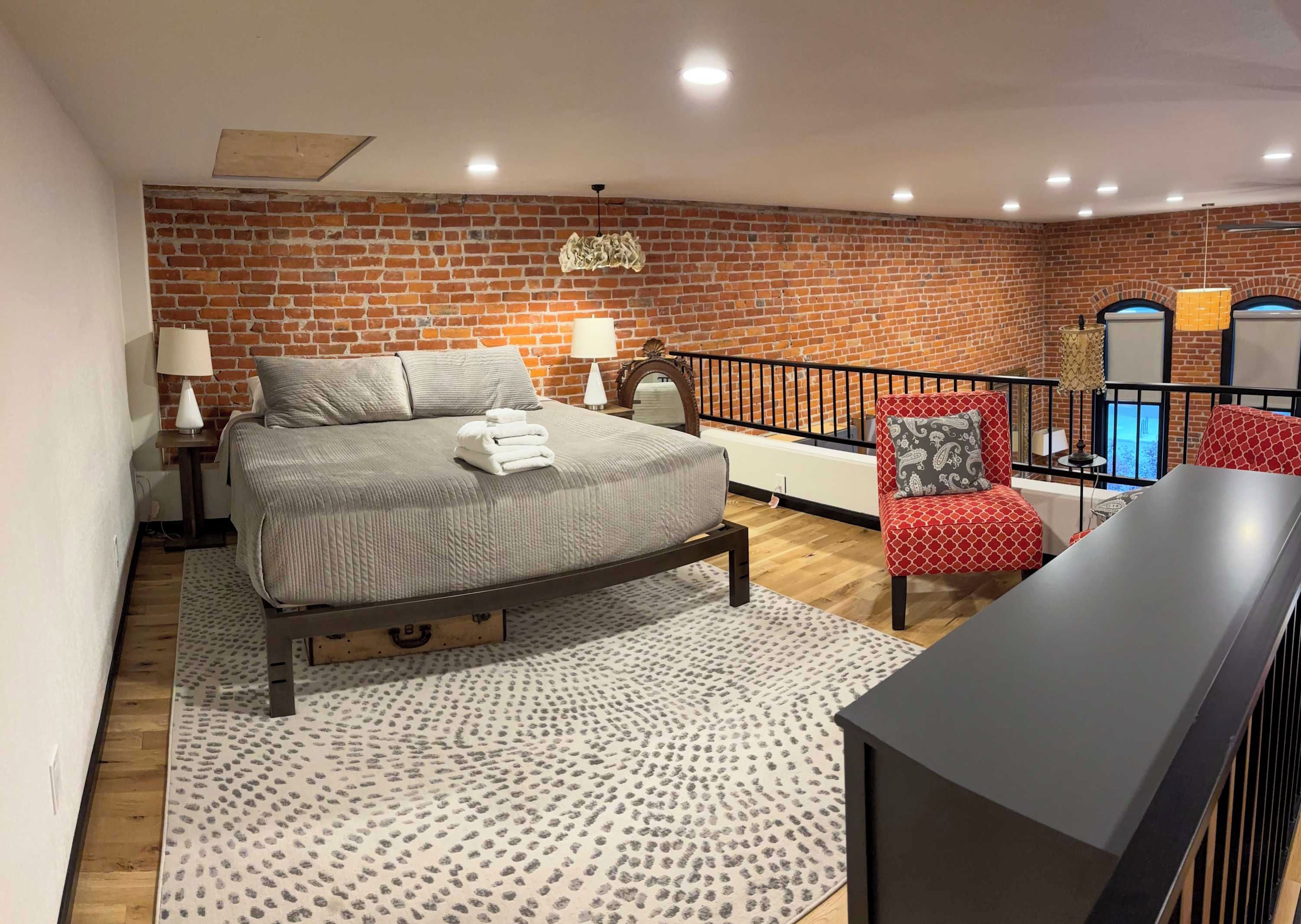 A bedroom with a brick wall, a bed with a headboard, a nightstand, a lamp, a red patterned chair, a black table, and a balcony with a railing.
