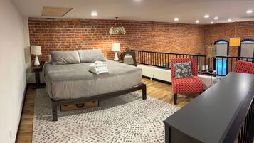 A bedroom with a brick wall, a bed with a headboard, a nightstand, a lamp, a red patterned chair, a black table, and a balcony with a railing.