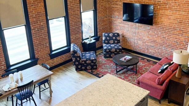 A room with a brick wall, a flat-screen TV mounted on the wall, a dining table with chairs, a sofa, and a small rug. - Luxury Downtown Loft w/ Panoramic Views & (Baker City)