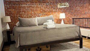 A bedroom with a brick wall, a bed with a wooden frame, and a nightstand with a lamp. - Luxury Downtown Loft w/ Panoramic Views & (Baker City)