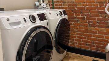 A front-loading washer and dryer set against a brick wall, with a wooden floor and a vent system above the dryer.