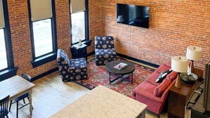 A room with a brick wall, a flat-screen TV mounted above a sofa, a dining table with chairs, and a patterned rug. - Luxury Downtown Loft w/ Panoramic Views & (Baker City)