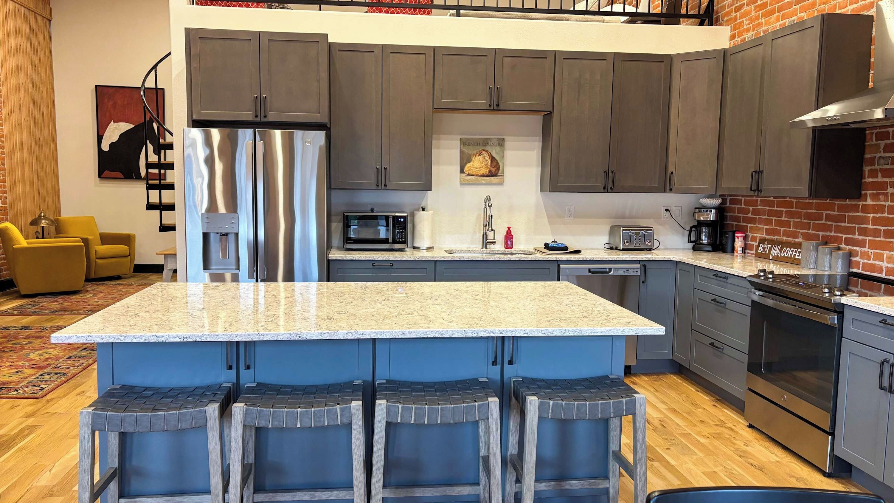 A modern kitchen with a central island, grey cabinets, stainless steel appliances, and a brick accent wall.