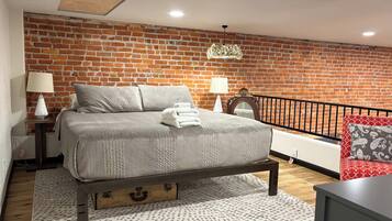 Een slaapkamer met een bakstenen muur, een bed met houten poten, twee bedlampjes, een rood kussen met een patroon en een klein tafeltje.