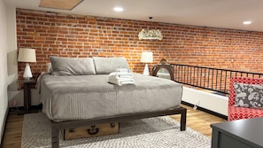 A bedroom with a brick wall, a bed with a wooden base, two bedside lamps, a red patterned pillow, and a small table. - Fishers Loft A , Luxury Downtown Loft w/ Panoramic Views (Baker City)