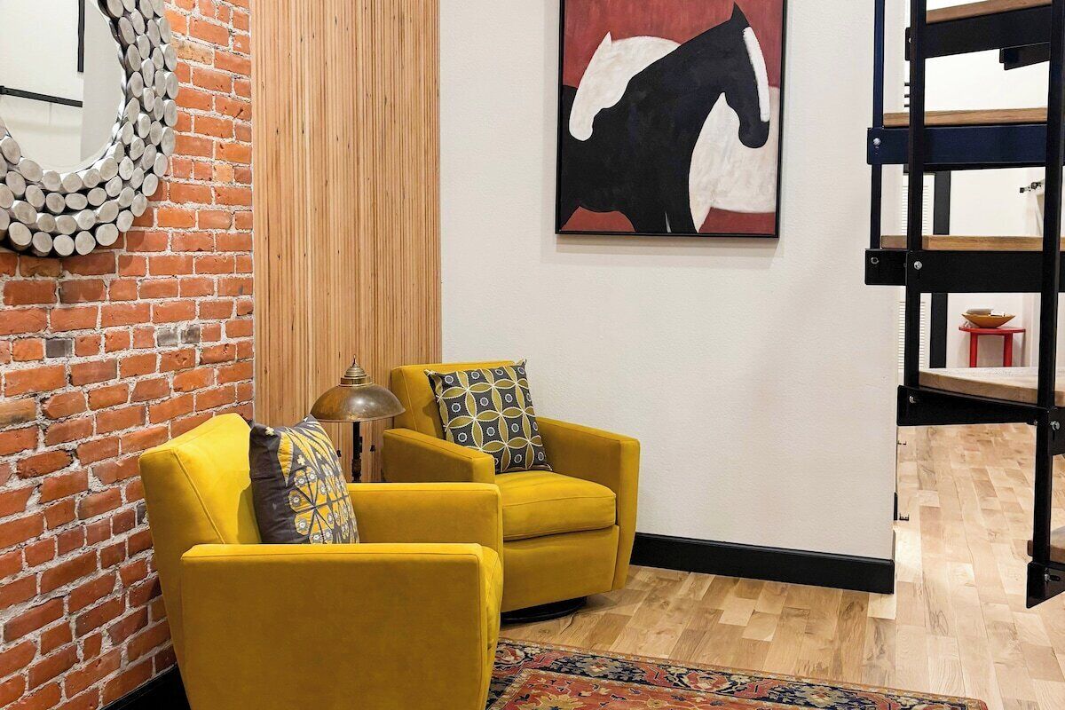 A room with a brick wall, a mirror, two yellow armchairs, a rug, a painting of a horse, and a wooden floor.