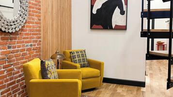 A room with a brick wall, a mirror, two yellow armchairs, a rug, a painting of a horse, and a wooden floor.