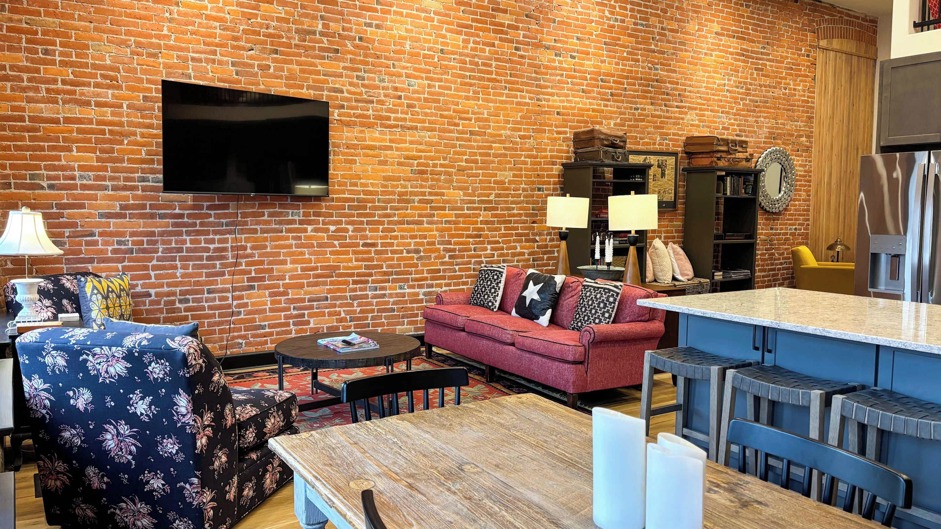 A living room with a brick wall, a flat-screen TV mounted above a sofa, a coffee table, and a dining area with a wooden table and chairs.