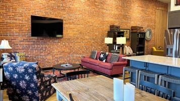 A living room with a brick wall, a flat-screen TV mounted above a sofa, a coffee table, and a dining area with a wooden table and chairs.