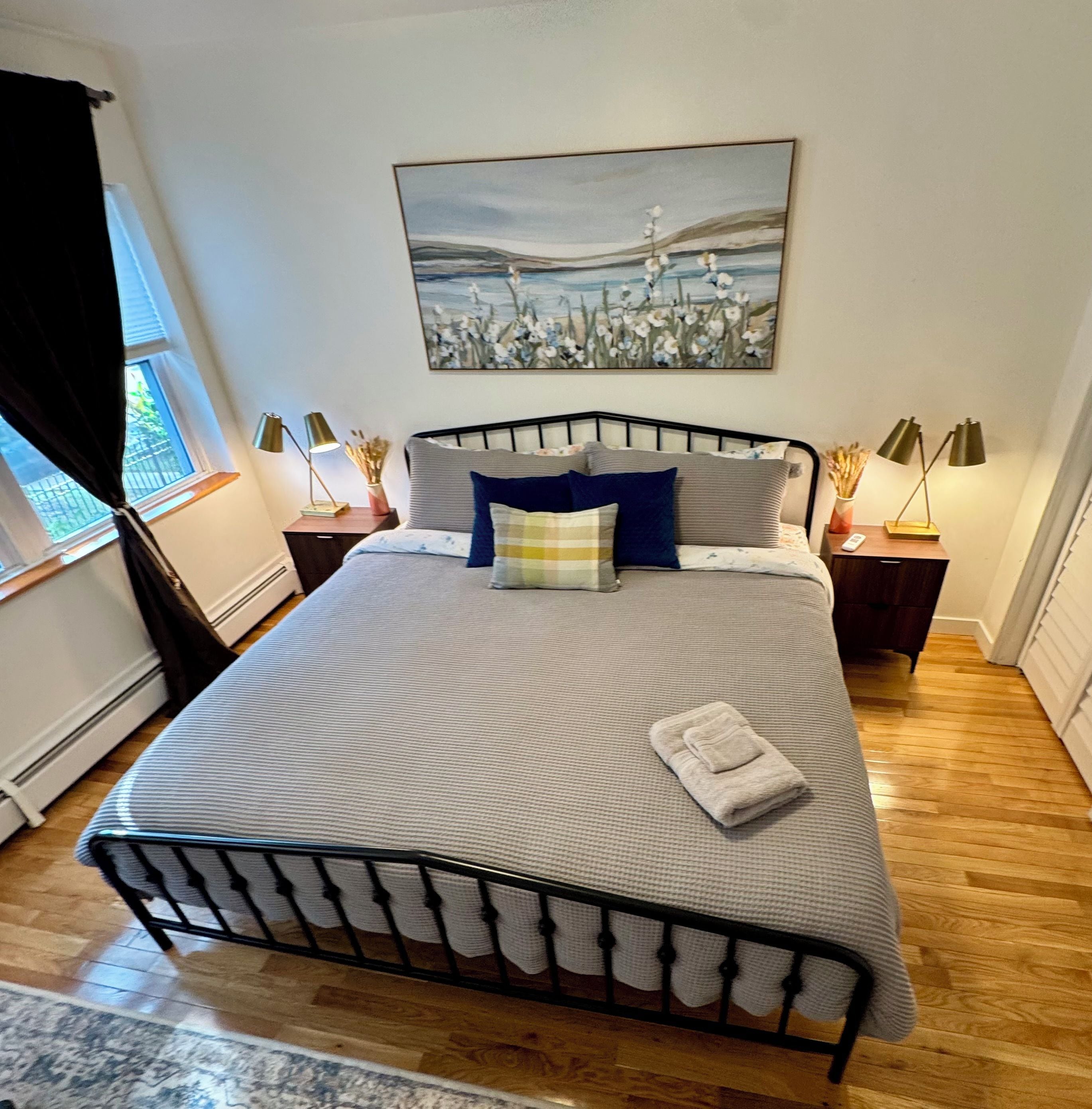 A neatly made bed with a patterned bedspread, a pillow, and a folded towel on a metal frame bed in a room with hardwood flooring.