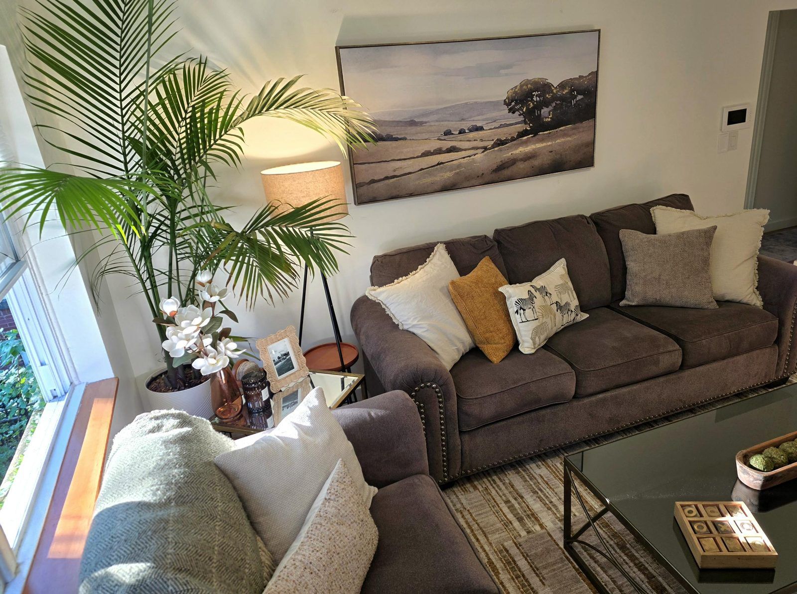 A living room with a brown sofa, a glass coffee table, a potted plant, and a painting on the wall.