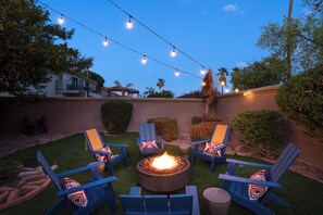 An outdoor seating area with blue chairs and a fire pit, surrounded by a well-maintained lawn and a house in the background. - Designer Home • Free Heated Pool • BBQ • Game Room • Garage • Sleeps 16 (Gilbert)