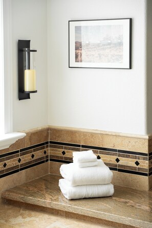 A bathroom with a marble countertop, a stack of white towels, a framed picture on the wall, and a wall-mounted light fixture. - Designer Home • Free Heated Pool • BBQ • Game Room • Garage • Sleeps 16 (Gilbert)