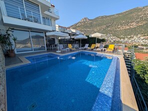 A swimming pool with lounge chairs and umbrellas, surrounded by a modern building with large windows. - Fantastic 3-bed Modern Villa with stunning views of Kalkan Bay from every level (Kalkan)