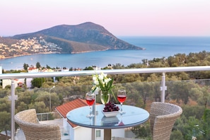 A balcony with a glass table, wicker chairs, a vase with flowers, and two glasses of red wine. - Fantastic 3-bed Modern Villa with stunning views of Kalkan Bay from every level (Kalkan)
