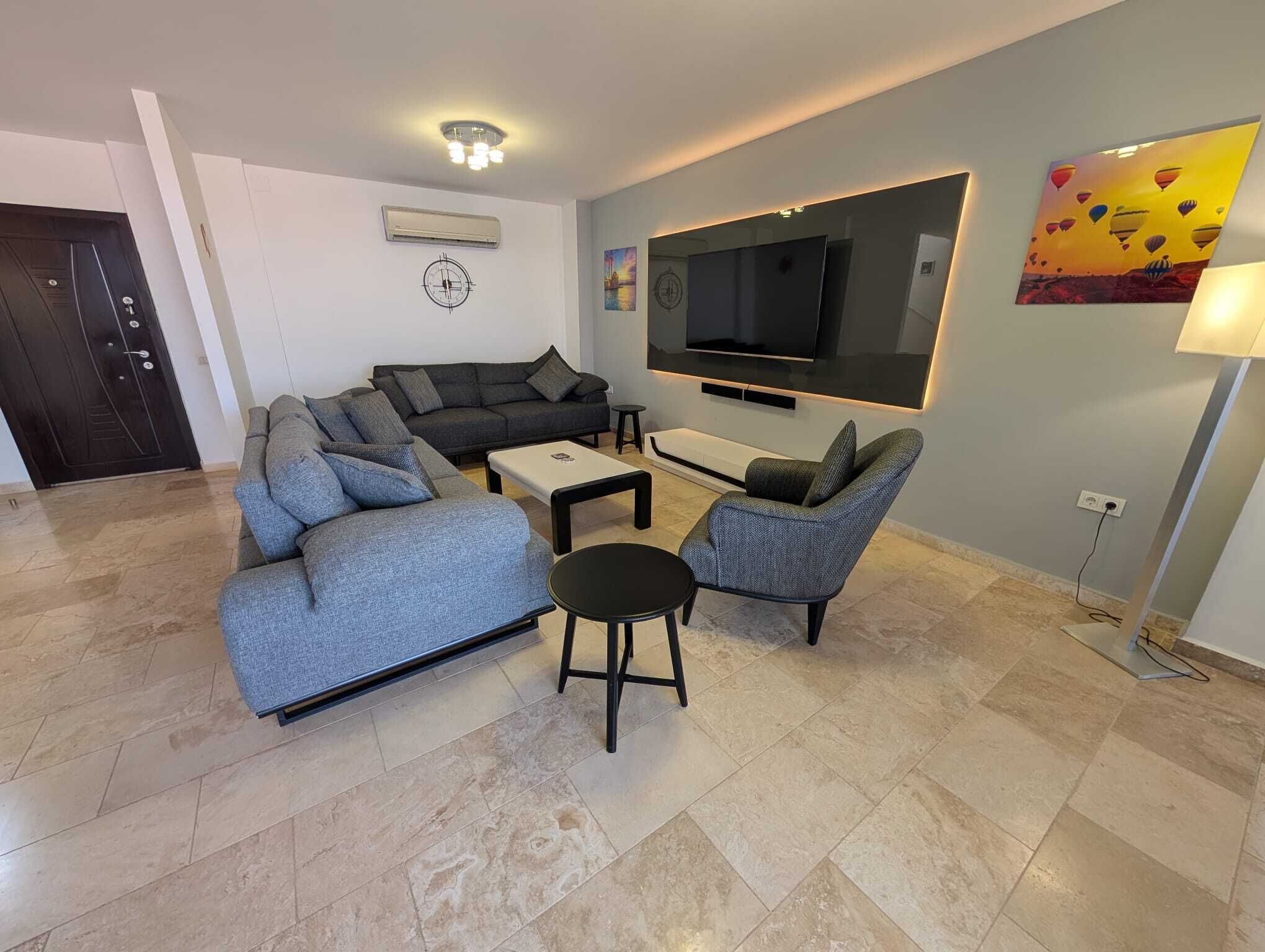 A modern living room with a sofa, armchair, coffee table, and a flat-screen TV mounted on the wall.