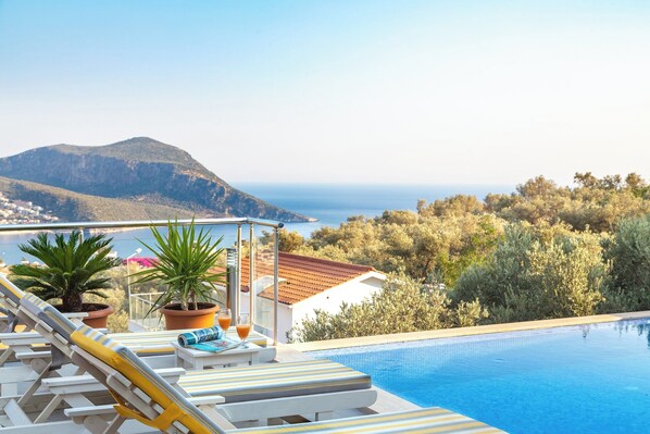 Ein Poolbereich mit Liegestühlen und einem Tisch, mit Blick auf eine Berglandschaft und das Meer.