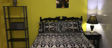 A bed with a patterned bedspread, a bedside lamp, a shelf with a vase of flowers, and a wall-mounted picture.