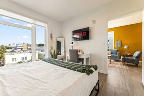 A hotel room with a bed, a desk, a chair, a TV, and a view of the city. - Bright & Chic 2BDR Suite K-Town Haven, Hot tub + Gym (Los Angeles)