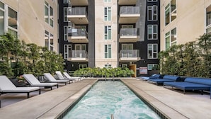 Une piscine entourée de chaises longues et d’un canapé bleu, le tout bordé par un immeuble doté de balcons.