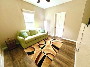 Una sala de estar con sofá, alfombra estampada, piso de madera, una puerta y un ventilador de techo.