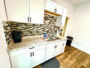 A compact kitchen with white cabinets, a granite countertop, a tiled backsplash, a double sink, and a coffee maker. - Massive on Myrtle St - 7 mins from DT - Sleeps 15 (Jacksonville)