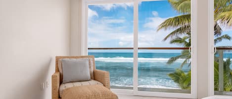 A room with a wicker chair and ottoman, a large sliding glass door to the ocean, and a view of palm trees.