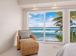 A room with a wicker chair and ottoman, a large sliding glass door to the ocean, and a view of palm trees. - Beachfront North Shore Oahu Estate | Sunset Beach (Haleiwa)
