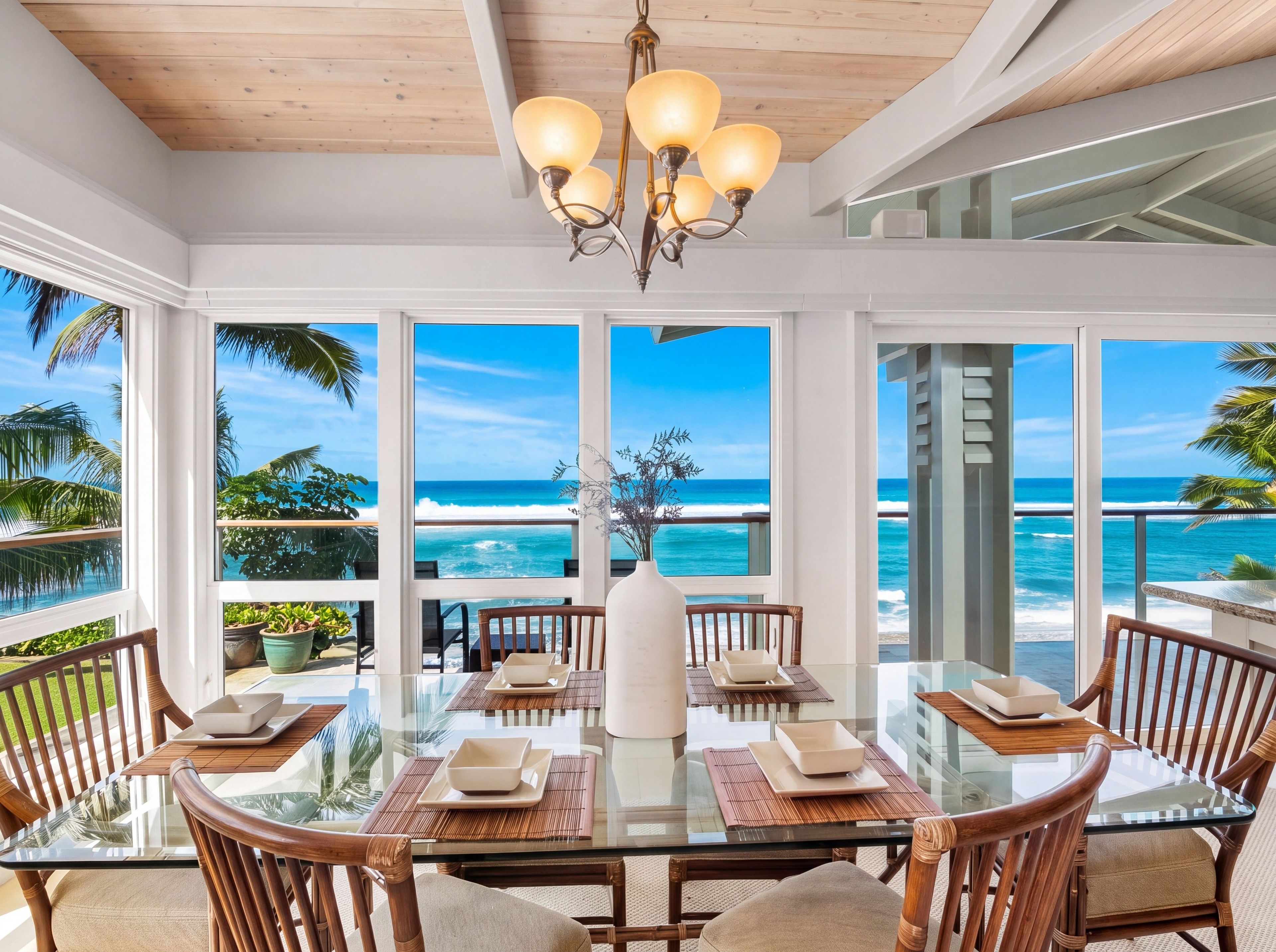A dining area with a glass table and chairs, a white vase centerpiece, and a view of the ocean.