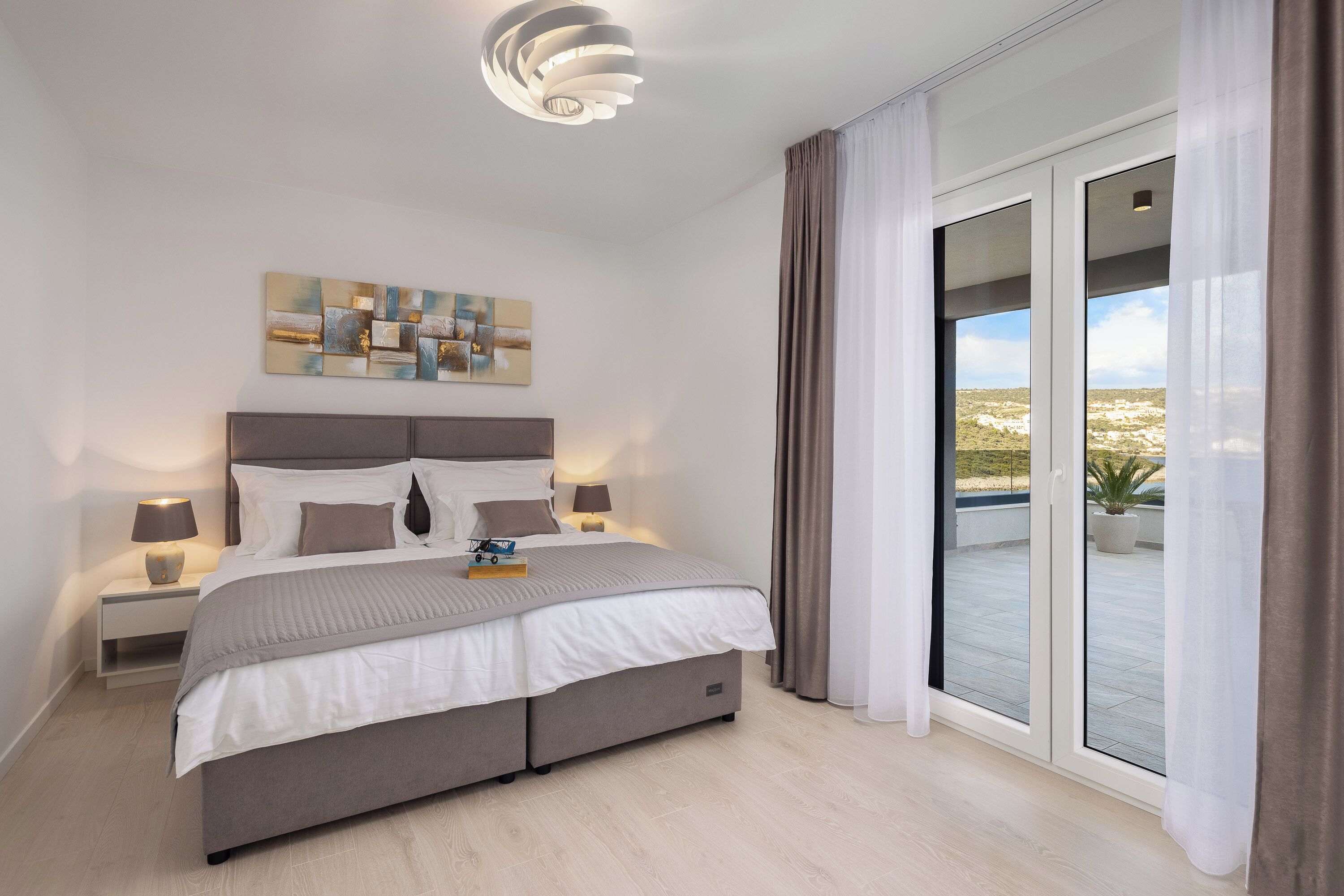 Une chambre à coucher moderne avec un grand lit, des lampes de chevet, un luminaire au plafond, une vue à travers une porte-fenêtre coulissante et une œuvre d’art abstraite accrochée au mur.