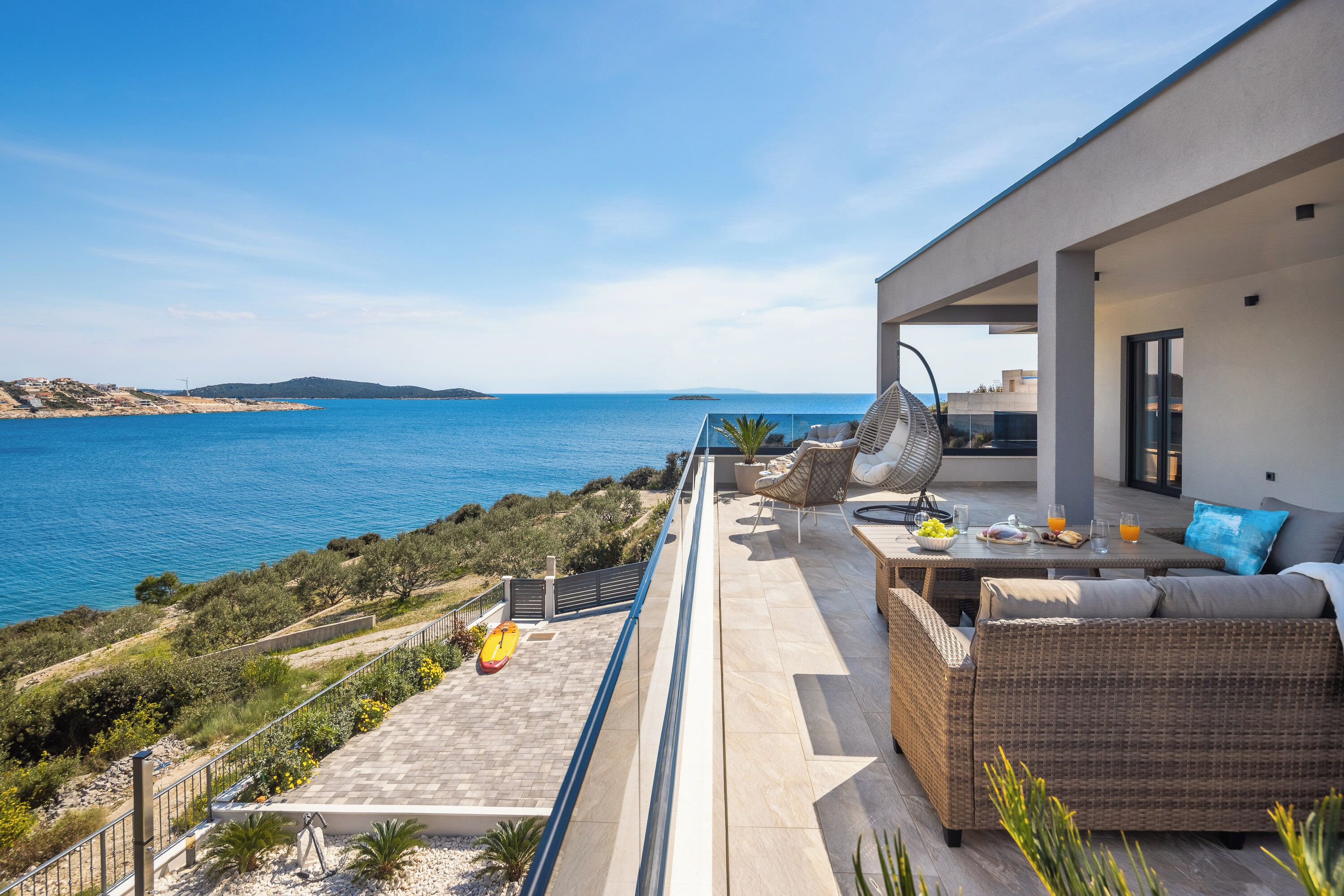 A modern outdoor terrace with a view of the sea, furnished with wicker furniture and a dining table.