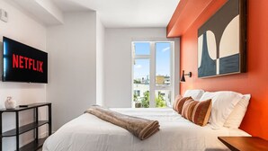 A modern hotel room with a large bed, a flat-screen TV mounted on the wall, a window with a city view, and a painting above the bed. - Chic 2 room suite in K-Town, Free Parking, Hot tub + Gym (Los Angeles)