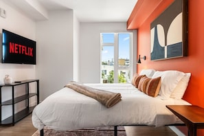 Ett modernt hotellrum med en stor säng, en platt-TV som är monterad på väggen, ett fönster med utsikt över staden och en tavla ovanför sängen.