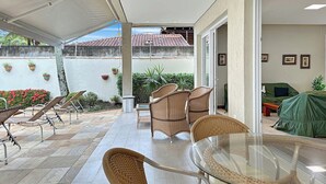 Área de varanda externa com móveis de vime, mesa de vidro e vista para um edifício com telhado em azulejos.