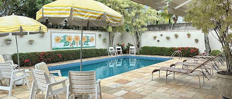 Zona piscina con lettini, ombrelloni a righe e piante in vaso.
