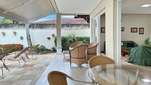Una veranda riparata all'aperto con mobili in vimini, un tavolo in vetro e vista su un edificio con tetto in tegole.