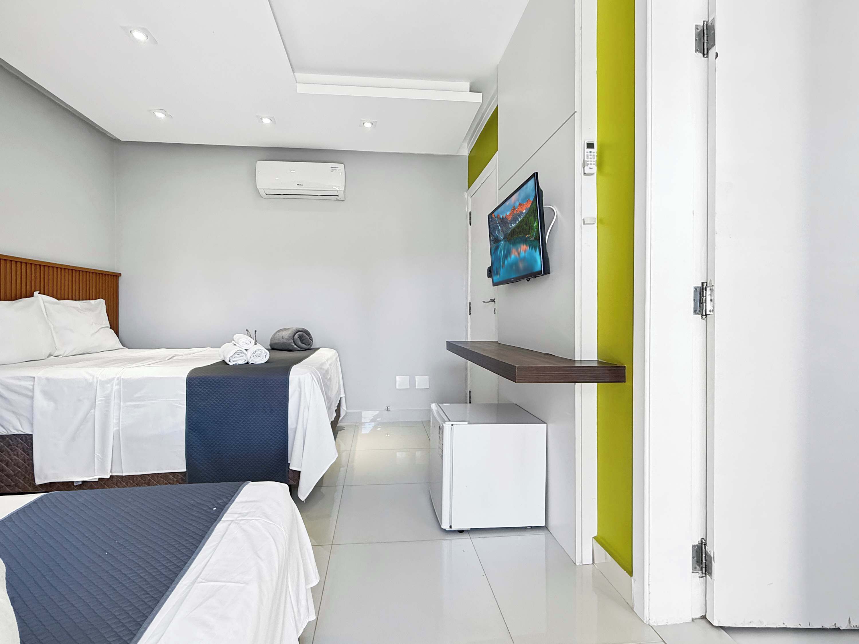 A hotel room with two beds, a TV mounted on the wall, a small white cabinet, and a door with a green accent.